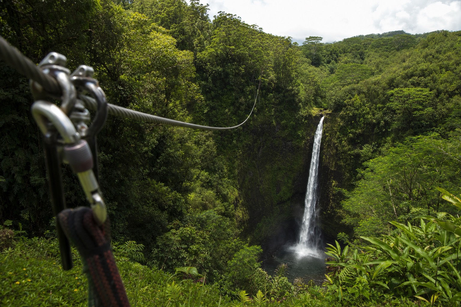Zipline cable stretching across a green Hamakua gulch toward a waterfall, with harness hardware in the foreground